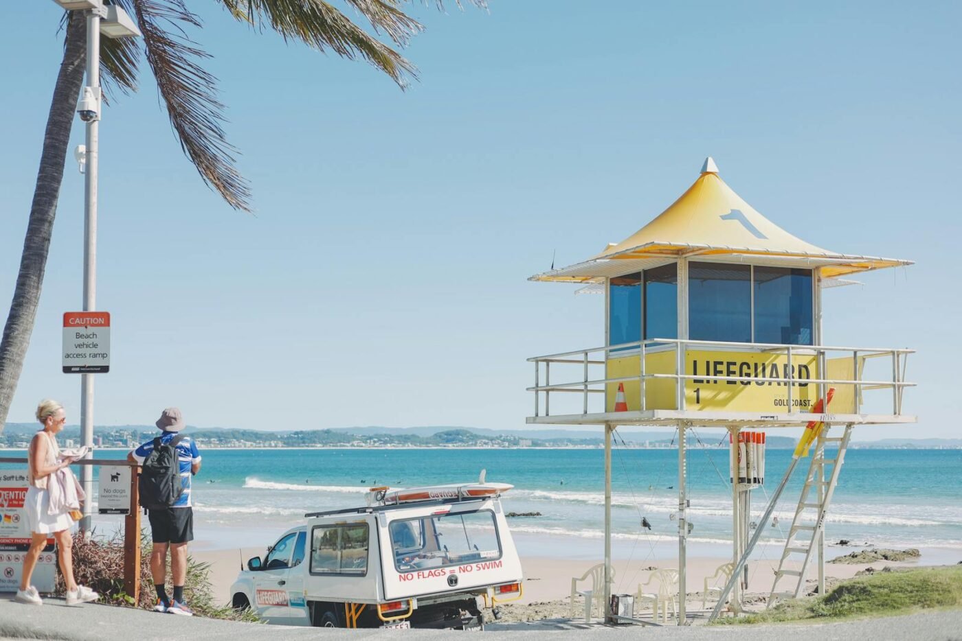 Sunny day at Coolangatta Beach with a lifeguard tower, palm tree, and people enjoying the shores.